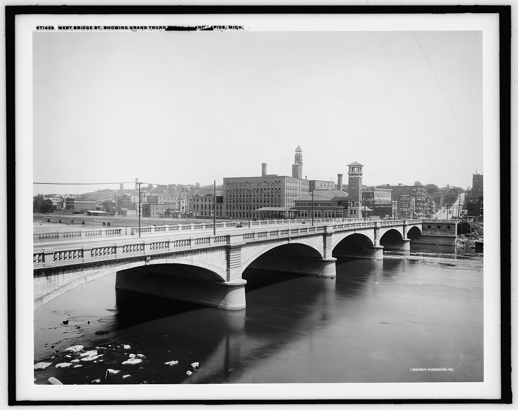 The Bridge Street bridge over the Grand River at Grand Rapids, photographed between 1900 and 1910, with the downtown skyline behind it and the water lying flat